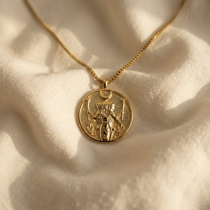 A gold Hecate triple goddess coin necklace with detailed medallion pendant and box chain lying flat on a white background with soft natural shadows, showing the intricate triple goddess symbolism, protective imagery, and polished gold finish from above.