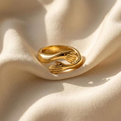 A gold hug ring with two detailed hands wrapping around to form an embrace lying flat on a white background with soft natural shadows, showing the intimate anthropomorphic design, textured fingers and palms, and polished gold finish from above.