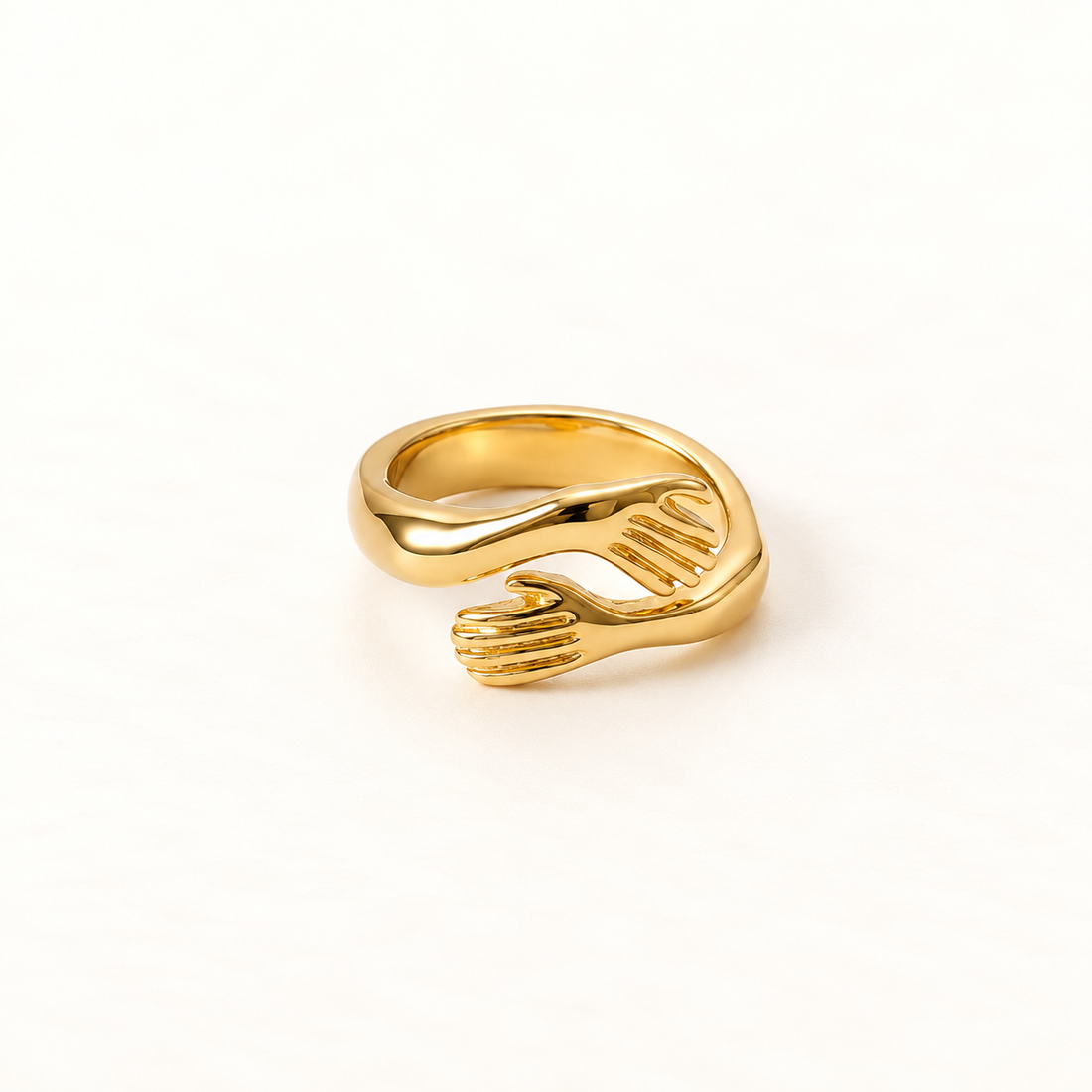 A gold hug ring with two detailed hands wrapping around to form an embrace, open adjustable band with textured fingers and palms, shown from front and side angles on a clean white background displaying the intimate anthropomorphic design, sculptural hand detail, and polished gold finish.