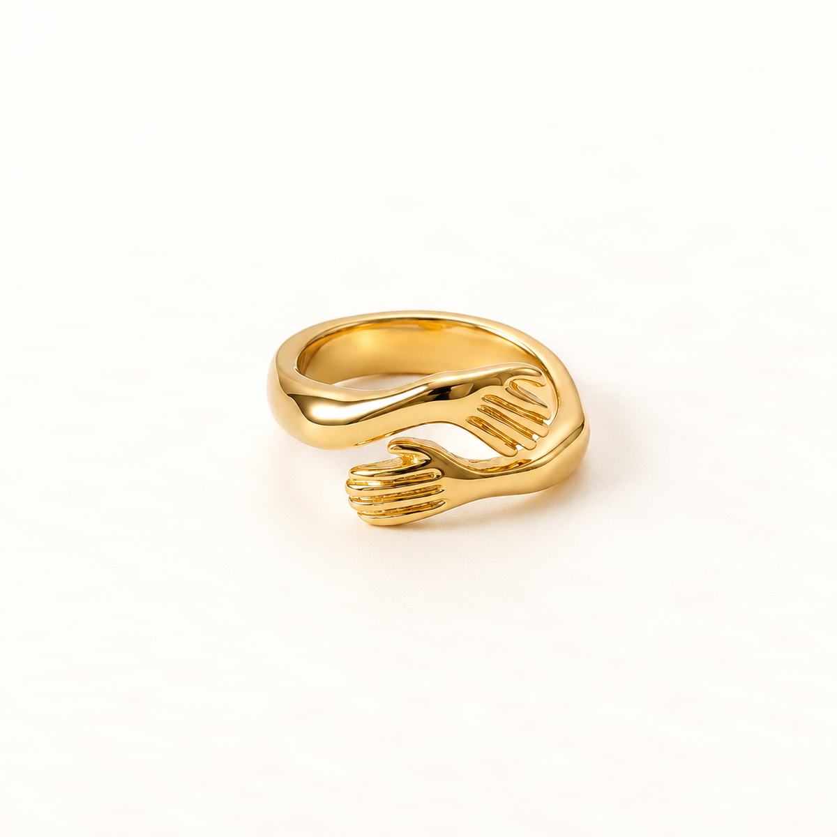 A gold hug ring with two detailed hands wrapping around to form an embrace, open adjustable band with textured fingers and palms, shown from front and side angles on a clean white background displaying the intimate anthropomorphic design, sculptural hand detail, and polished gold finish.