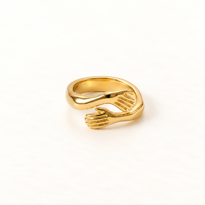 A gold hug ring with two detailed hands wrapping around to form an embrace, open adjustable band with textured fingers and palms, shown from front and side angles on a clean white background displaying the intimate anthropomorphic design, sculptural hand detail, and polished gold finish.