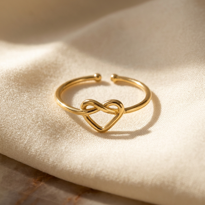 A gold infinity heart ring with continuous loop forming an intertwined heart shape lying flat on a white background with soft natural shadows, showing the eternal love symbolism, delicate interwoven design, and polished gold finish from above.