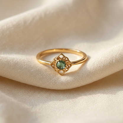 A gold jade filigree ring with green cabochon center stone in diamond-shaped ornate setting lying flat on a white background with soft natural shadows, showing the rich green hue, intricate lace-like frame, and polished gold finish from above.