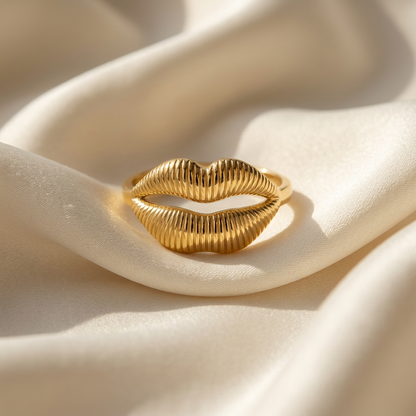 A gold lips kiss ring with detailed puckered mouth design and textured lip lines lying flat on a white background with soft natural shadows, showing the sculptural mouth form, intricate lip texture, and polished gold finish from above.