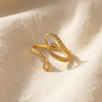 A gold octopus tentacle ring with detailed suction cup texture and curved tentacle arms lying flat on a white background with soft natural shadows, showing the intricate marine texture, organic flowing form, and polished gold finish from above.