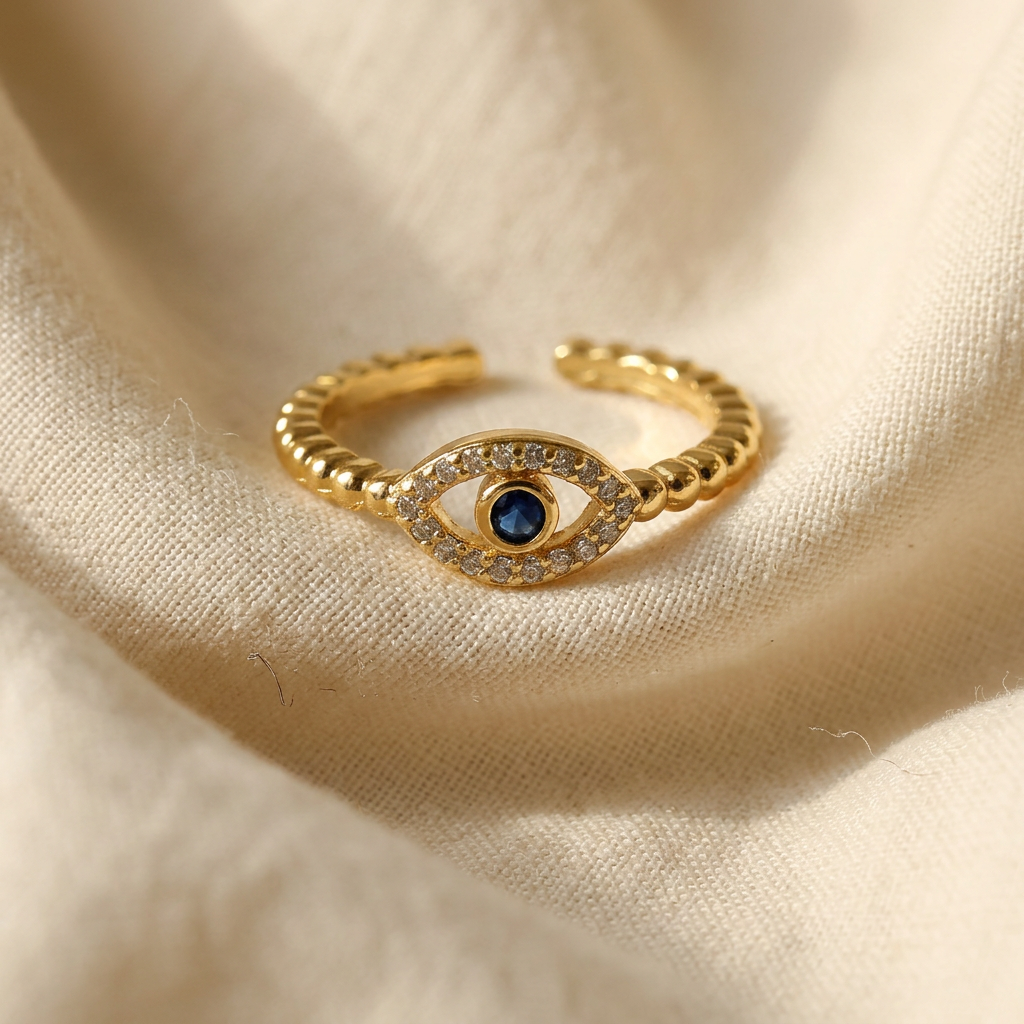 A gold pave evil eye ring with beaded ball chain band, clear crystals surrounding deep blue center stone lying flat on a white background with soft natural shadows, showing the protective symbol, sparkling stone detail, and polished gold finish from above.
