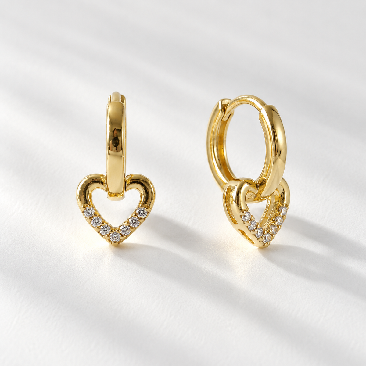 Two 18k gold heart-shaped huggie hoop earrings with clear pave crystals lying on a white background with soft natural shadows, one showing the front sparkling detail and one displaying the hinged huggie closure mechanism.