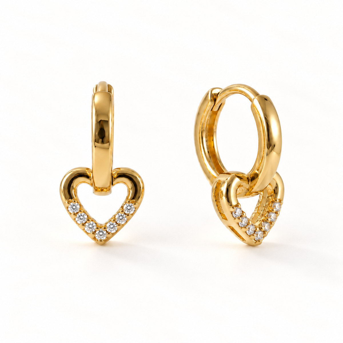 A pair of 18k gold heart-shaped huggie hoop earrings with clear pave crystals along the edges, shown from front and side angles on a clean white background displaying the hinged closure and sparkling stone detail.