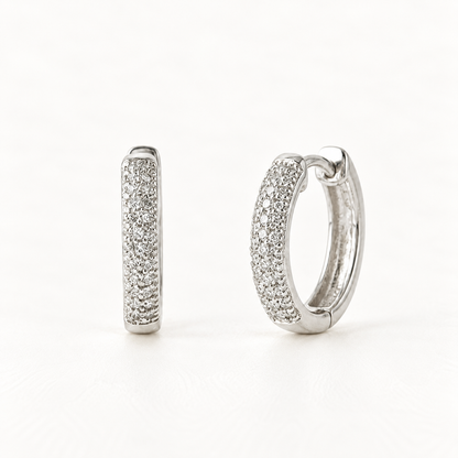 A pair of sterling silver pave huggie hoop earrings with clear crystals set along the front half, shown from front and side angles on a clean white background displaying the sparkling stone detail and hinged closure.