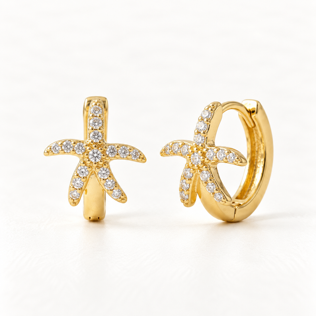 A pair of 18k gold starfish-shaped huggie hoop earrings with clear pave crystals covering the surface, shown from front and side angles on a clean white background displaying the sparkling five-point sea-inspired detail and hinged closure.