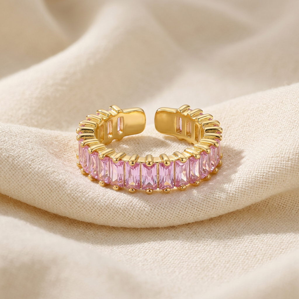 A gold pink baguette ring with continuous band of rectangular pink crystals lying flat on a white background with soft natural shadows, showing the soft rose hue, sparkling step-cut stones, and polished gold finish from above.