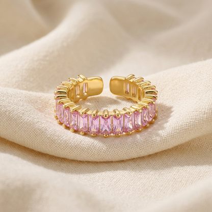 A gold pink baguette ring with continuous band of rectangular pink crystals lying flat on a white background with soft natural shadows, showing the soft rose hue, sparkling step-cut stones, and polished gold finish from above.
