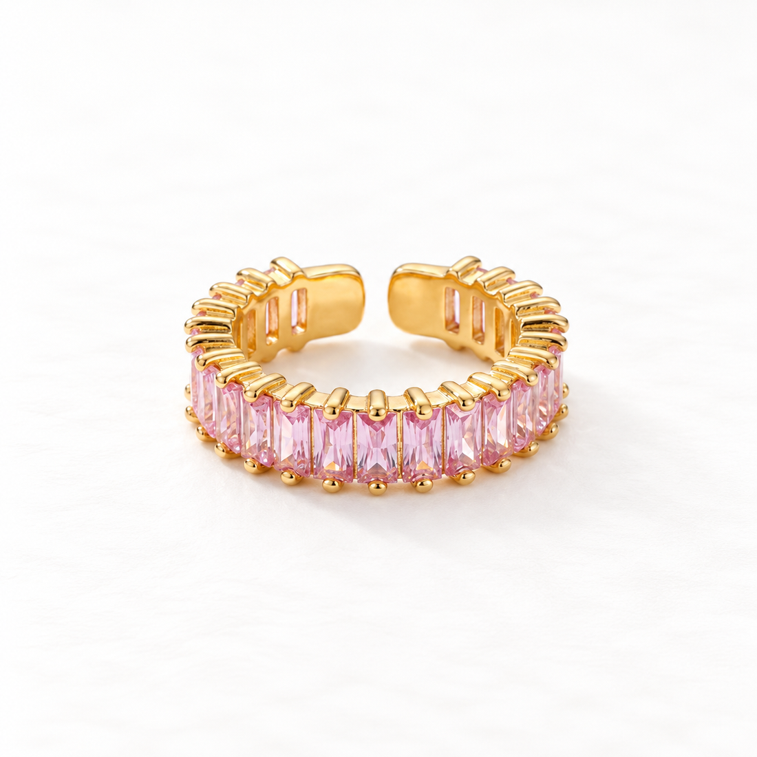 A gold pink baguette ring with continuous band of rectangular pink crystals, eternity-style open adjustable design with prong settings, shown from front and side angles on a clean white background displaying the soft rose hue, sparkling step-cut stones, and polished gold finish.