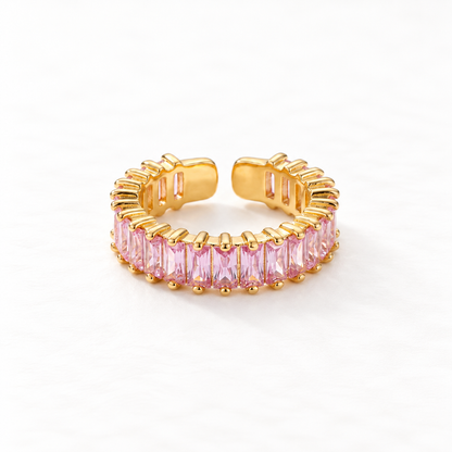 A gold pink baguette ring with continuous band of rectangular pink crystals, eternity-style open adjustable design with prong settings, shown from front and side angles on a clean white background displaying the soft rose hue, sparkling step-cut stones, and polished gold finish.
