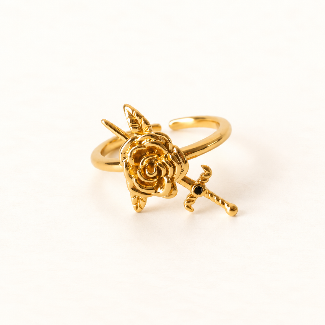 A gold rose and dagger ring with detailed blooming rose and crossed sword design, open adjustable band, shown from front and side angles on a clean white background displaying the romantic yet edgy symbolism, intricate petal and blade texture, and polished gold finish.