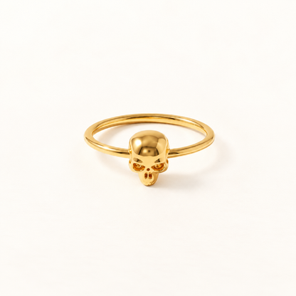 A gold skull ring with delicate band and small detailed skull centerpiece featuring hollow eye sockets and defined jawline, shown from front and side angles on a clean white background displaying the minimalist band, edgy yet feminine aesthetic, and polished gold finish.
