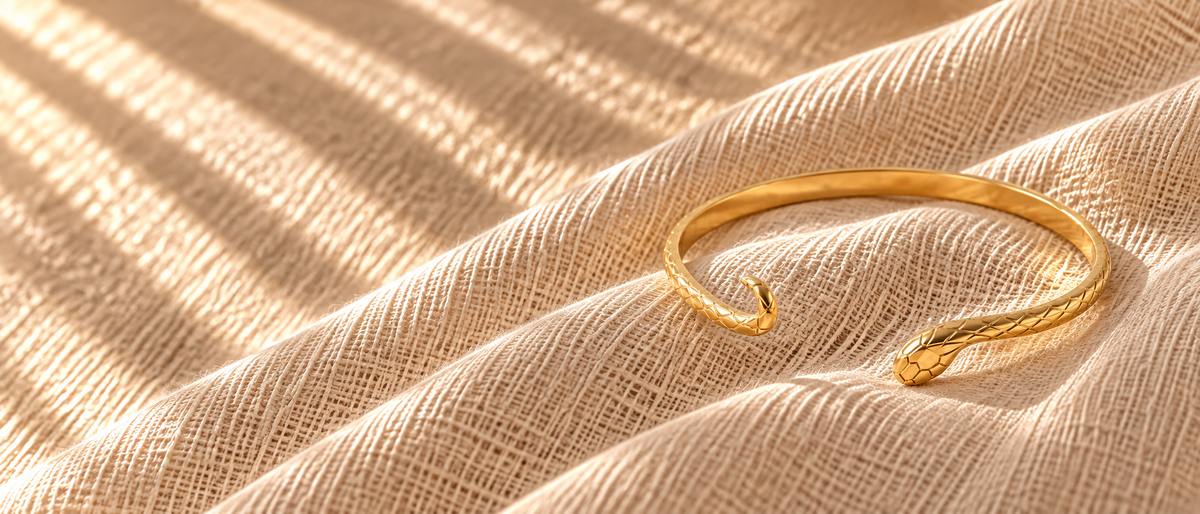 Gold snake bangle bracelet on oatmeal linen fabric with dappled sunlight creating soft shadows. Textured scale pattern and serpent head detail visible. Warm golden tones, organic fabric texture, intimate jewelry photography.