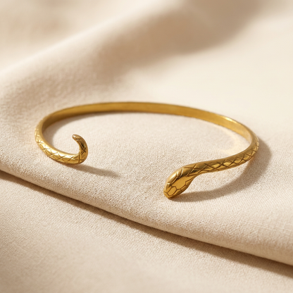 A gold snake cuff bracelet with textured scale pattern and detailed serpent head lying flat on a white background with soft natural shadows, showing the open cuff design, curved tail end, and intricate scale texture from above.