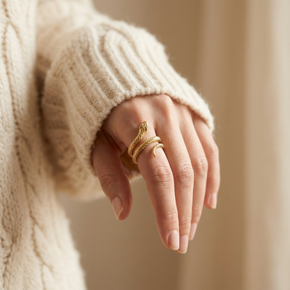 Close-up of a feminine hand wearing a gold snake wrap ring with detailed scale texture, the coiled serpent body wrapping elegantly around the finger with head and tail visible. Soft natural lighting, warm beige linen background, natural skin texture with visible pores and slight imperfections.
