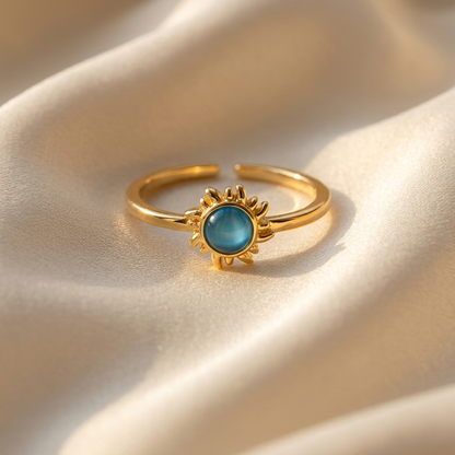 A gold sun ring with vibrant blue turquoise center stone and radiating sunburst rays lying flat on a white background with soft natural shadows, showing the bright blue hue, detailed ray texture, and polished gold finish from above.