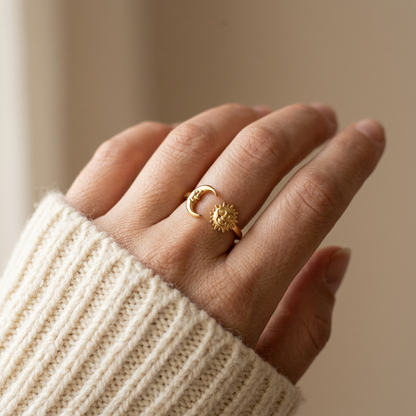 Close-up of a feminine hand wearing a gold sun and moon open ring with anthropomorphic sun face and crescent moon charms, the celestial symbols resting on the finger. Soft natural lighting, warm beige linen background, natural skin texture with visible pores and slight imperfections.