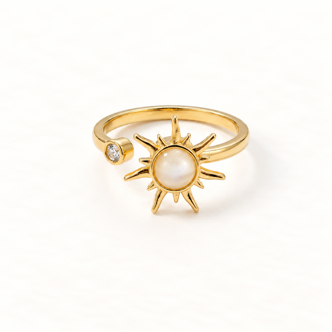 A gold sun spinner ring with rotating sunburst center, pearlescent moonstone center and small clear crystal accent, open adjustable band, shown from front and side angles on a clean white background displaying the kinetic celestial design, radiating rays, and polished gold finish.