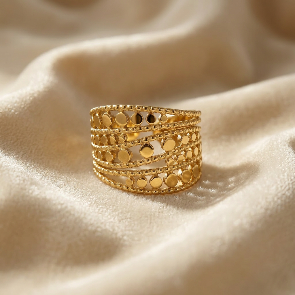 A gold wide band cutout ring with multiple rows of circular geometric cutouts and beaded edge details lying flat on a white background with soft natural shadows, showing the intricate lattice pattern, substantial presence, and polished gold finish from above.