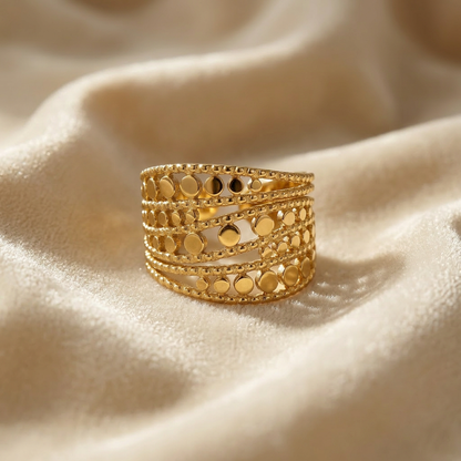 A gold wide band cutout ring with multiple rows of circular geometric cutouts and beaded edge details lying flat on a white background with soft natural shadows, showing the intricate lattice pattern, substantial presence, and polished gold finish from above.