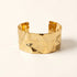 A wide hammered gold cuff bracelet with substantial bold presence, textured dimpled surface, and irregular organic edges, shown from front and side angles on a clean white background displaying the artisan hammered detail, sculptural form, and polished gold finish.